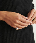 A person in a black ribbed sweater gently clasps their hands, showcasing the shiny Felix Dome Ring in white gold on their pinky finger.
