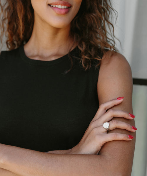 A woman with wavy hair, pink lipstick, and red nail polish crosses her arms, displaying the stunning Gemma Baguette Signet Ring in 14k gold on her finger. She pairs it with a sleeveless black top.