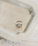 The Gemma Baguette Signet Ring—a white gold ring with a square-cut gemstone—rests on a rectangular, light metal tray featuring raised edges and a slightly weathered look.