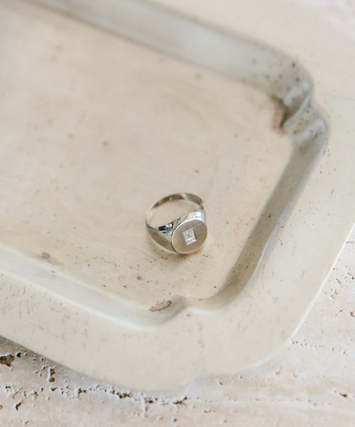 The Gemma Baguette Signet Ring—a white gold ring with a square-cut gemstone—rests on a rectangular, light metal tray featuring raised edges and a slightly weathered look.