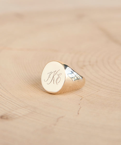 The Gemma Signet Ring in 14k gold features engraved cursive initials JKC and is displayed on a light wooden surface with natural wood grain.