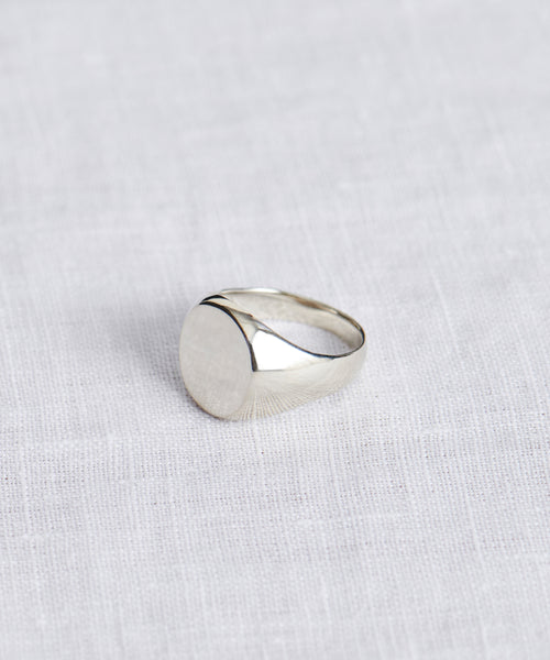The Gemma Signet Ring, featuring a plain silver band with a round, flat top, is shown on a white textured fabric background.