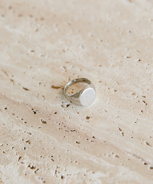 The Gemma Signet Ring, crafted in white gold with a plain round face, lies on a beige textured stone—an understated piece from Jenni Kayne Jewelry.