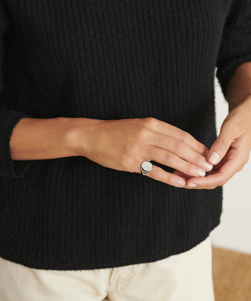 A person wears a black knit sweater and light pants, displaying manicured nails and the Gemma Signet Ring on one finger.