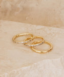 Three gold rings, including the Iris Eternity Band, are stacked on a smooth beige stone. The background features softly blurred neutral tones.