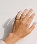 A light-skinned hand, wearing the Iris Eternity Band with conflict-free diamonds and two other 14k solid gold rings—a thick signet and a thin band—rests on a white surface.