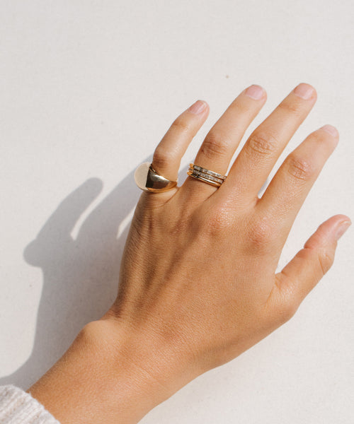 A light-skinned hand, wearing the Iris Eternity Band with conflict-free diamonds and two other 14k solid gold rings—a thick signet and a thin band—rests on a white surface.