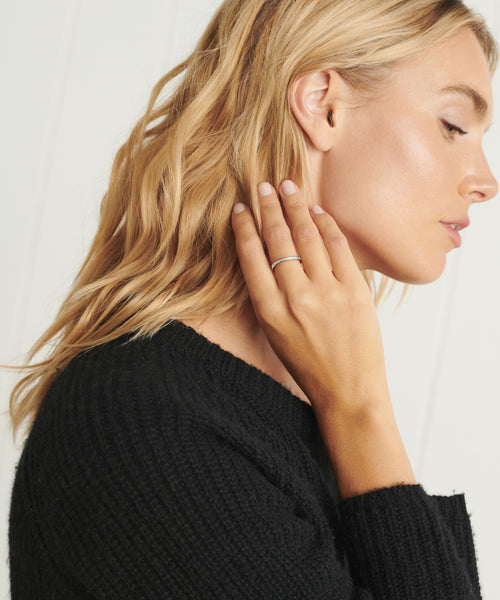 A blonde woman in a black sweater touches her neck with her left hand, highlighting the simple Iris Eternity Band on her finger as she faces sideways against a light background.