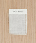 A rectangular Fabric Swatch Textured Natural Linen, made from Belgian flax linen, is displayed on a light wood surface with a card labeled jenni kayne in lowercase black letters attached at the top.