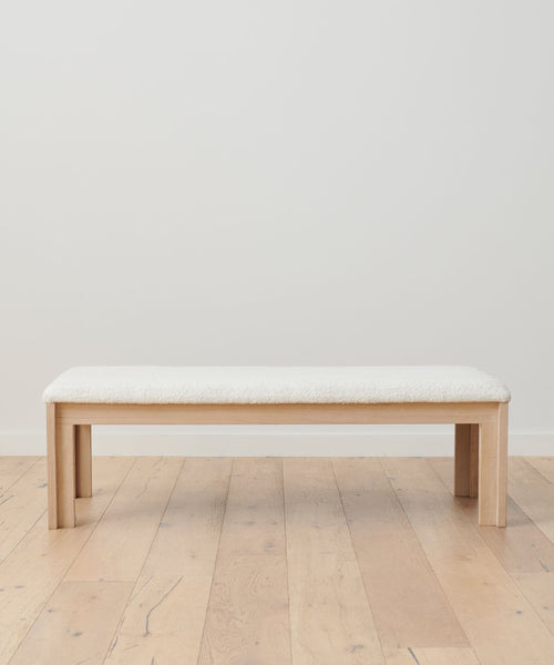 The Otto Bench showcases an Ivory Wool Boucle cushioned seat atop crisp White Oak, styled on a light wood floor with a plain white wall backdrop.