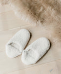 A pair of Shearling Moroccan Slippers with white fluffy shearling and soft suede outsoles rest on a light wooden floor beside a beige faux fur rug.