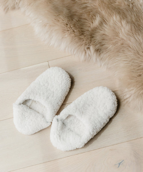 A pair of Shearling Moroccan Slippers with white fluffy shearling and soft suede outsoles rest on a light wooden floor beside a beige faux fur rug.