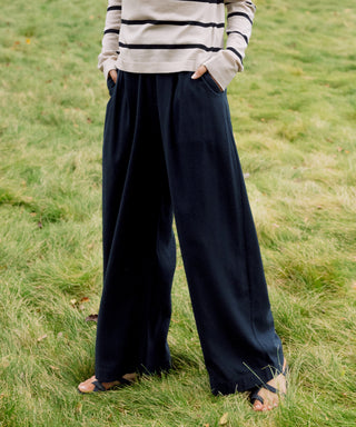 A person stands on grass wearing Relaxed Trouser wide-leg black pants, a beige and black striped long-sleeve top, and sandals. Their hands are in their pockets, and their face is not visible.