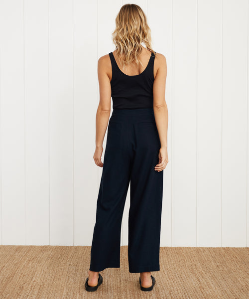 A woman with wavy blonde hair faces away, wearing a black sleeveless top and the Relaxed Trouser in a wide-leg style, paired with black shoes, set against a white paneled wall and woven rug.