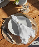 A table setting with a woven placemat, white plate, Ceramic Napkin Rings Set of 4 on a white cloth napkin, fork and knife, and a glass of water on a coaster atop a wooden California table. Sunlight casts shadows over the scene.