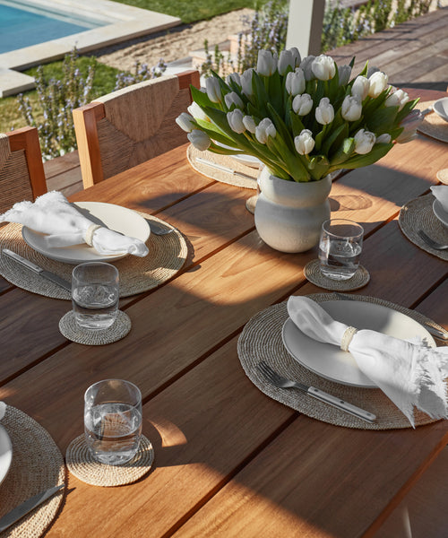 A handmade wooden outdoor dining table set with white plates, the Ceramic Napkin Rings Set of 4, glasses of water, woven placemats, and a vase of white tulips. A pool and lush California garden complete the inviting backdrop.