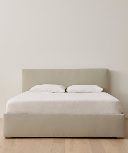 The Pacific Bed features a Belgian linen upholstered headboard and frame, neatly dressed in white sheets and pillows, positioned on a light wood floor against a plain white wall.
