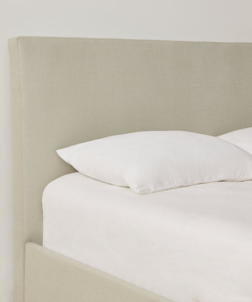 A close-up of the Pacific Bed features a light beige Belgian linen upholstered headboard, white pillow, and crisp white bedding set against a plain white wall.