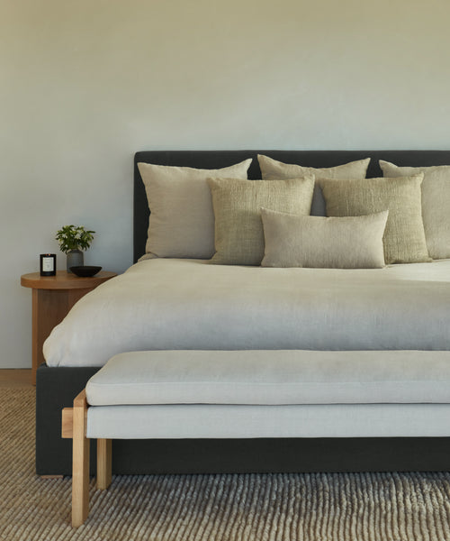 A modern bedroom with a neatly made Pacific Bed featuring a grey upholstered headboard, beige throw pillows, a light bench at the foot, a wooden nightstand with a small plant and candle, and a textured rug on the floor.
