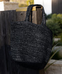 The Woven Raffia Tote in black hangs on a dark wooden fence outdoors, with greenery and a stone structure in the background.