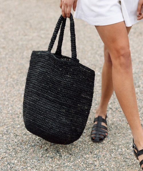 A person in white shorts and black sandals walks on gravel, carrying a large black Woven Raffia Tote by its handles.