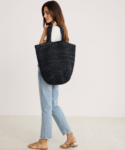 A woman in a white t-shirt, light blue jeans, and sandals stands in profile against a plain background with the Woven Raffia Tote slung over her shoulder.
